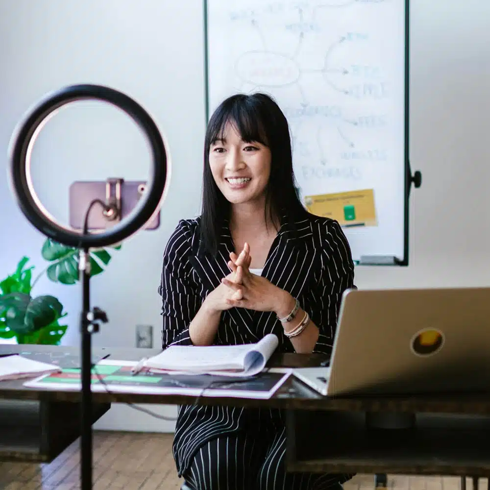 A smiling content creator records a video at her desk using a ring light and laptop, with notes and documents spread out in front of her and a whiteboard with a business diagram in the background.