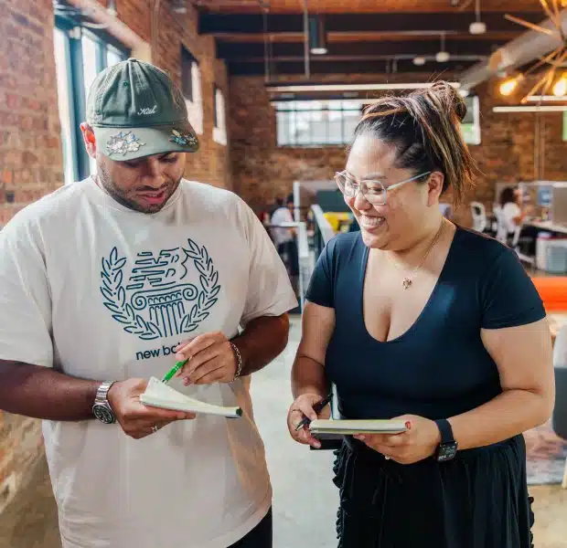 Ehab, a tax associate, and Darcil, a tax associate, stand together in a bright, modern office space with exposed brick walls, reviewing documents in their notebooks and smiling.